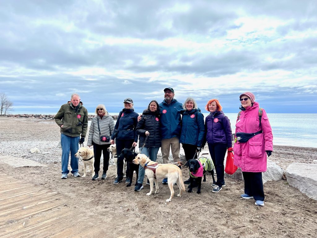 8 Staff and clients of Guide Dogs for the Blind stand on a beach with their dogs